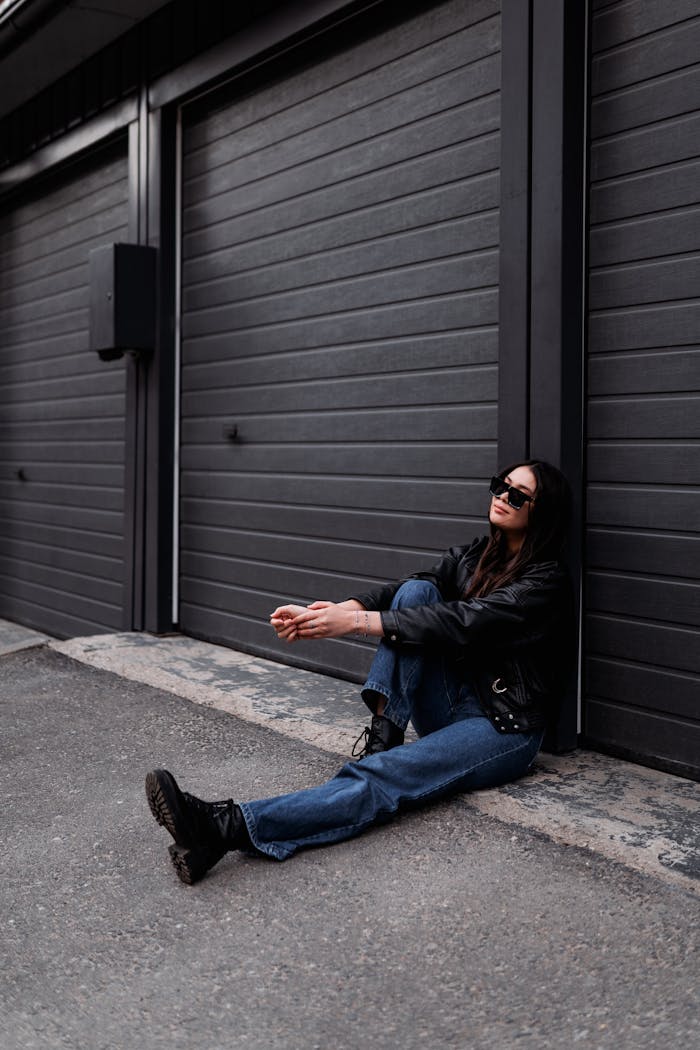 Stylish brunette woman in leather jacket and sunglasses sitting against garage doors.